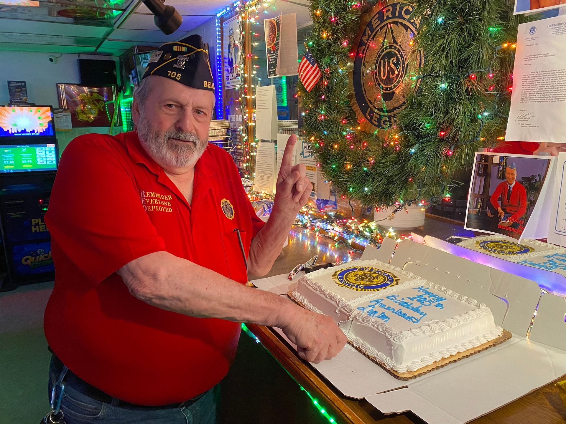 Man in red shirt points to cake, decorated with emblem, in a bar decorated for a veterans' event.