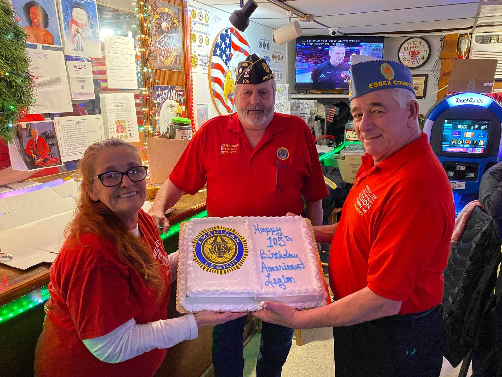 Three people hold a cake at an event. The cake has a logo and text. They wear red shirts.