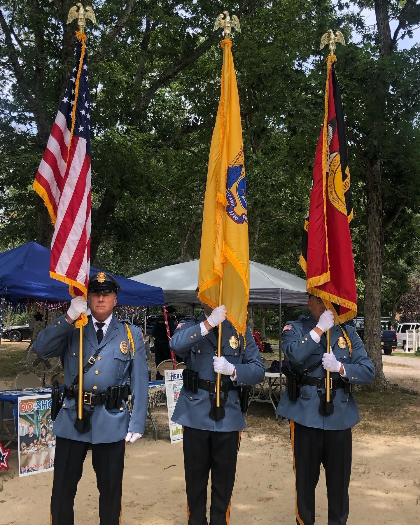 Three uniformed officers hold flags: American, yellow seal, and red crest, outdoors.