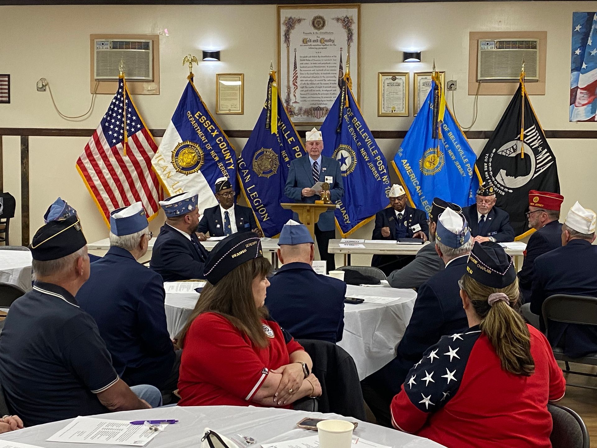 Veterans' meeting: speaker at podium, flags, people seated at tables, wearing hats, in a room.