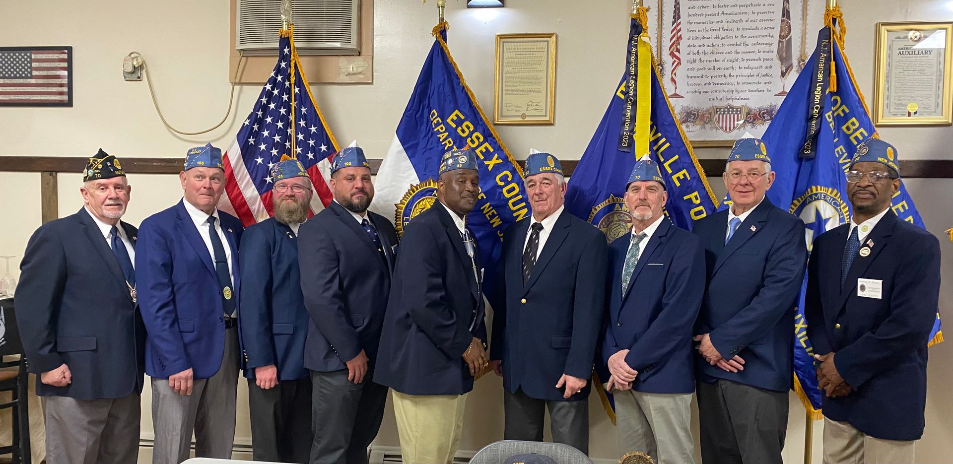 Group of men in uniform stand with flags. Some wear hats and suit jackets.