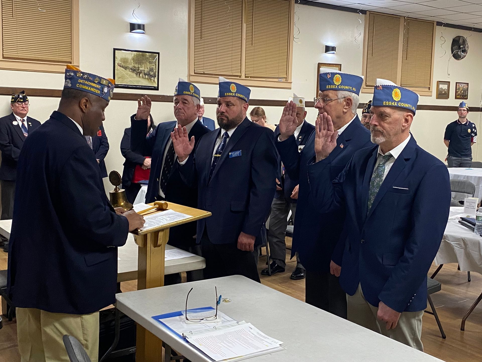 Men in blue blazers and hats take an oath with raised hands at a podium in a meeting hall.