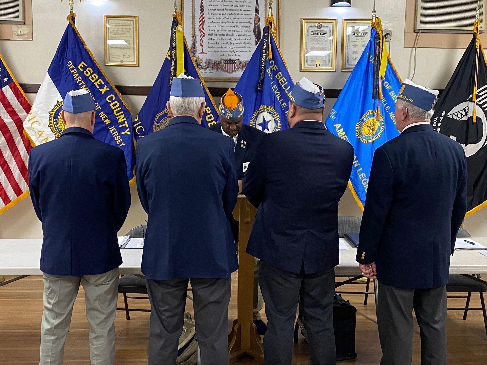 Four men in blue blazers and hats stand before flags in a room.