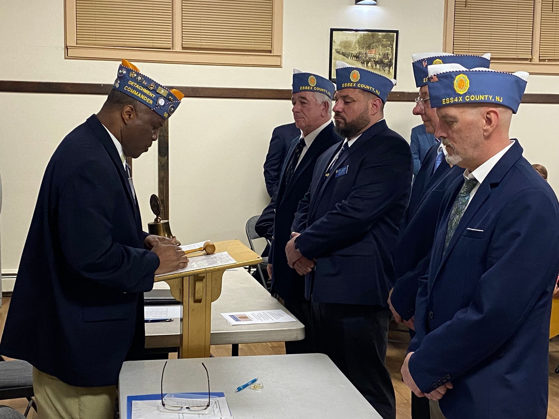 Men in navy blazers and caps at a lectern, observing a ceremony in a wooden-paneled room.