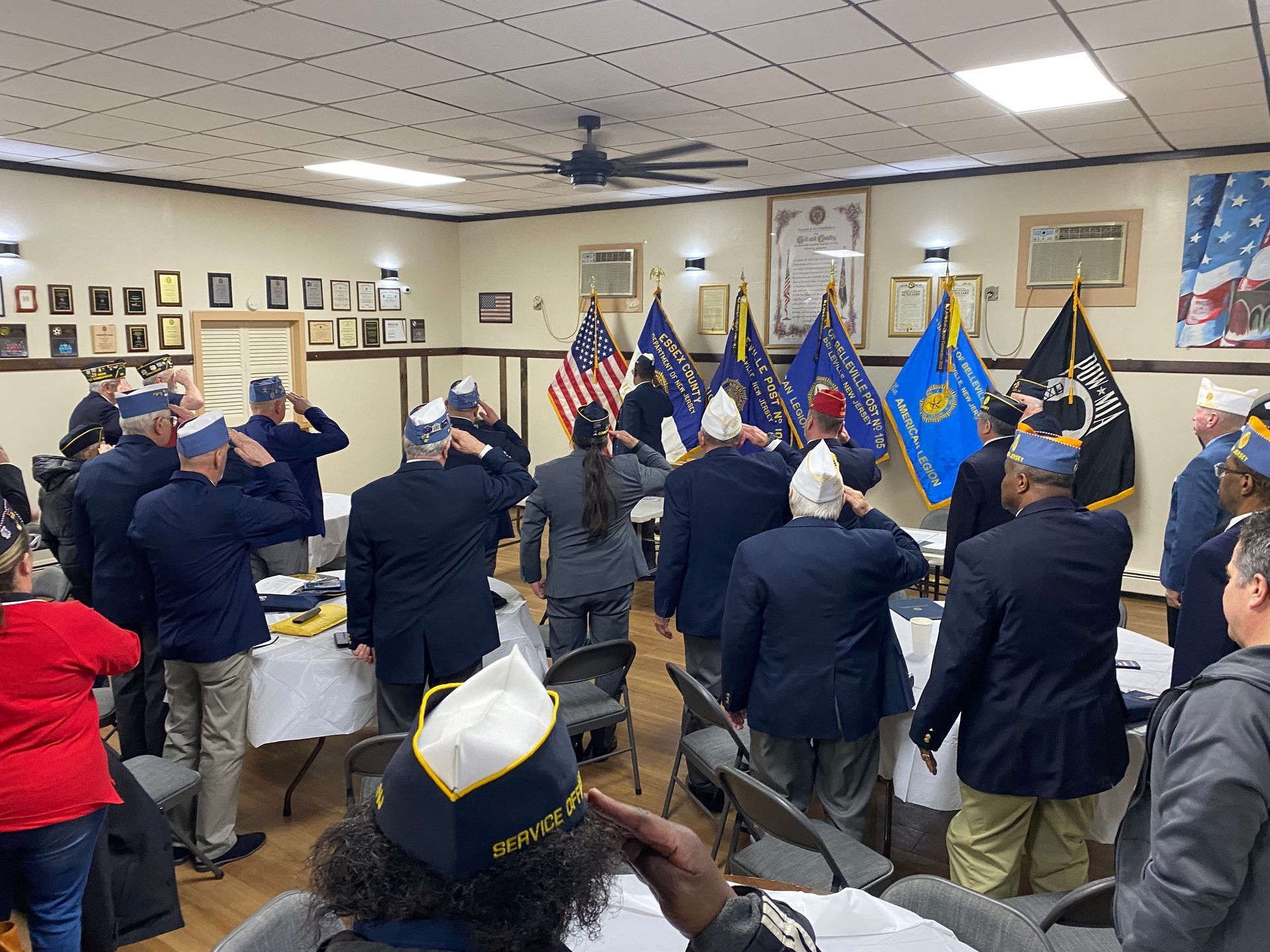 Veterans saluting indoors, American flags visible.  Men and women in blue coats and hats.