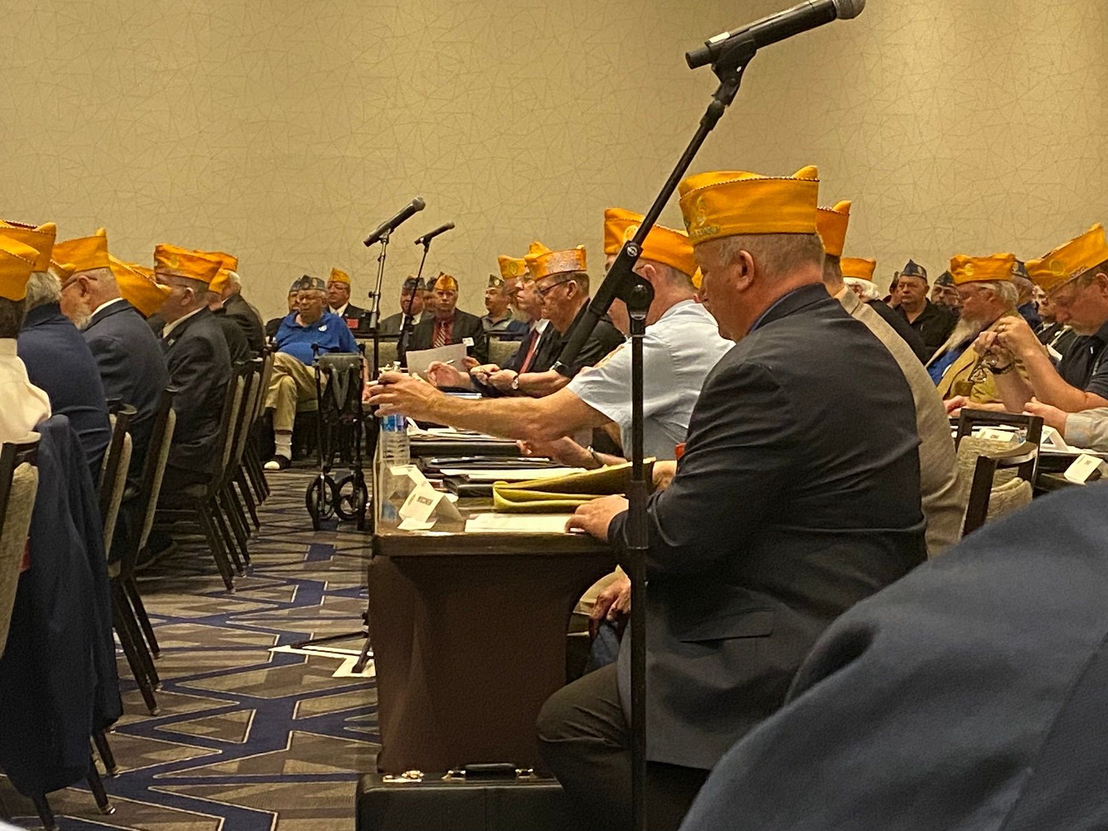 A meeting of veterans wearing yellow hats. They sit at tables with microphones, focused on the speaker.