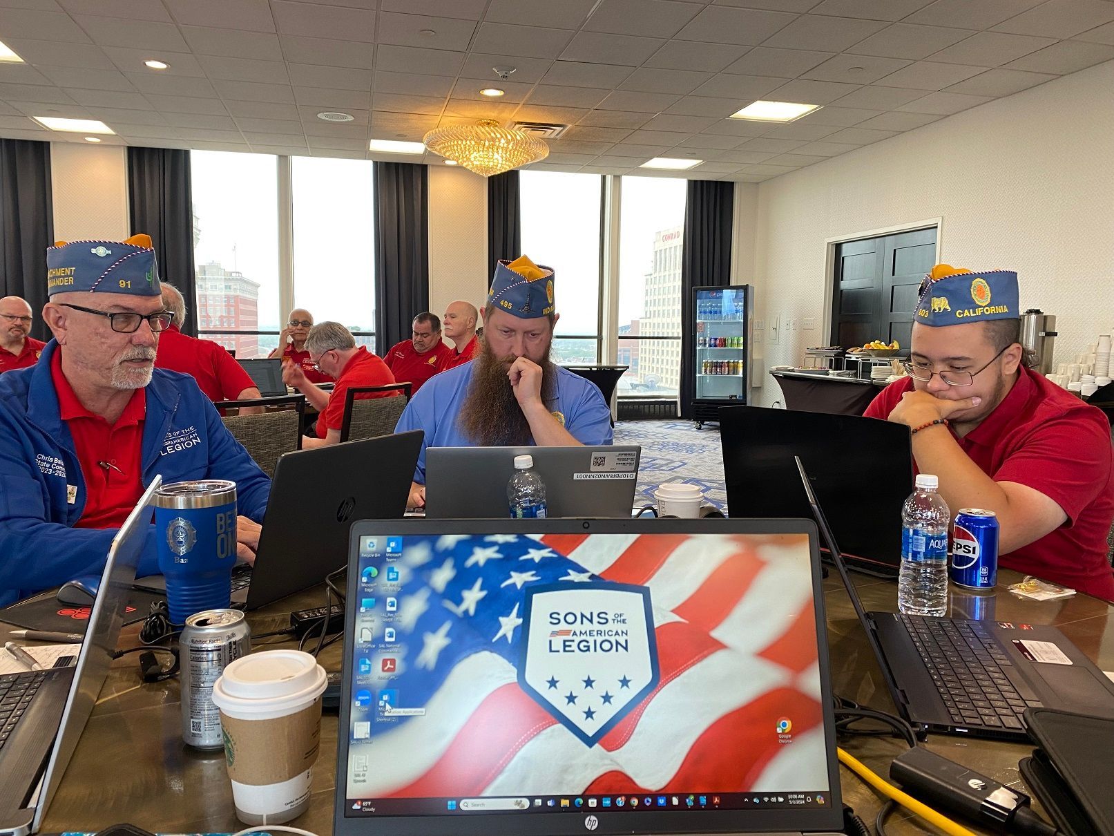 Men in blue and red attire with laptops, some wearing caps, in a meeting room, American flag background.