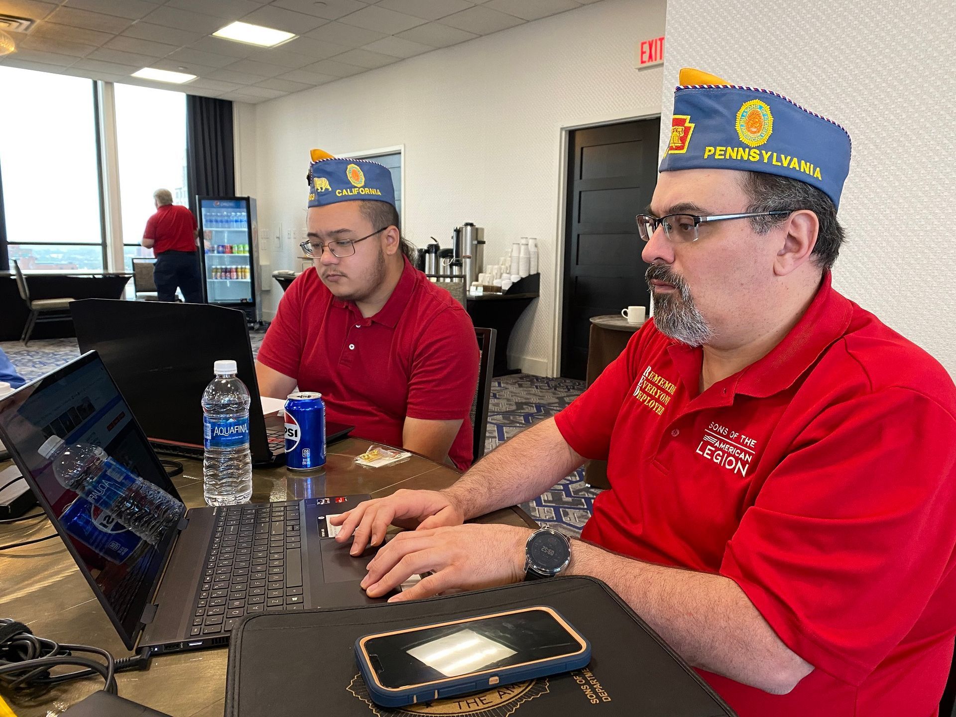 Two men in red shirts and blue hats with insignia, working at a laptop in a room.