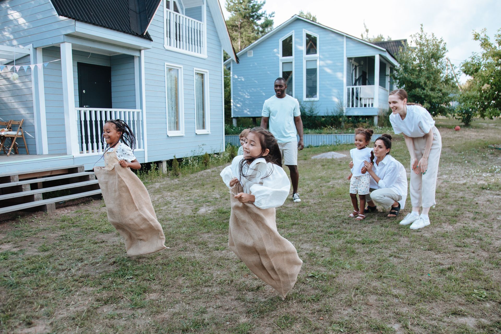 Children in burlap sacks race in a yard, adults watch nearby, blue houses in background.