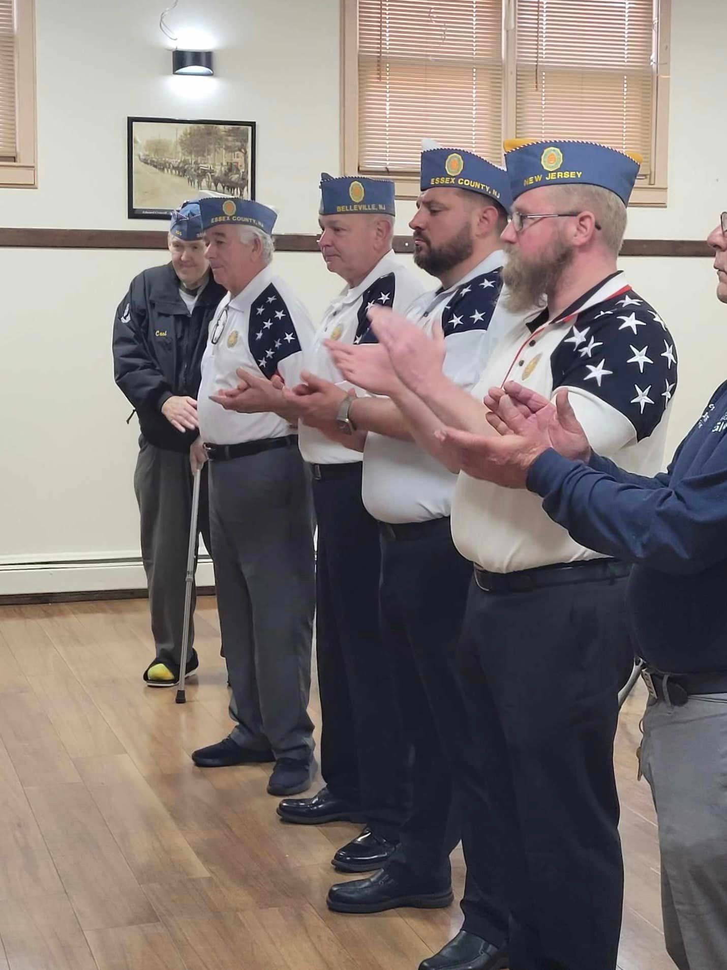Men in military-style uniforms applaud in a room with wood floors and a framed picture.