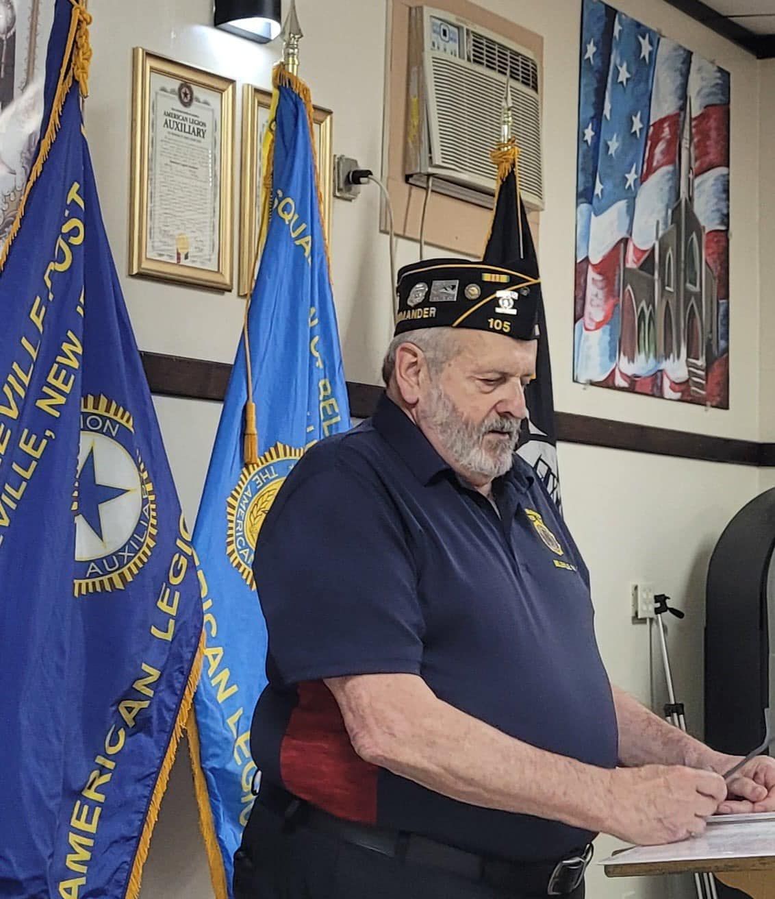 Man in navy shirt and hat with insignia, standing near blue flags.