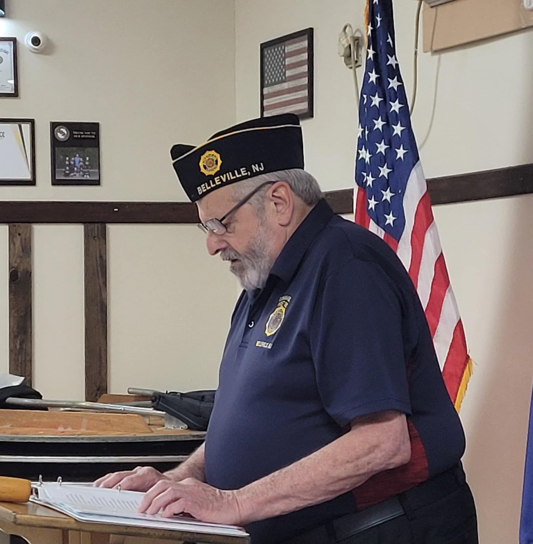 Man in veteran's hat reading at a table with an American flag behind him.