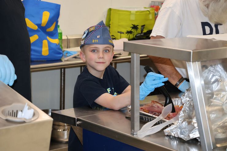 Boy in a wizard hat smiles while serving food at a counter, someone in gloves helps.