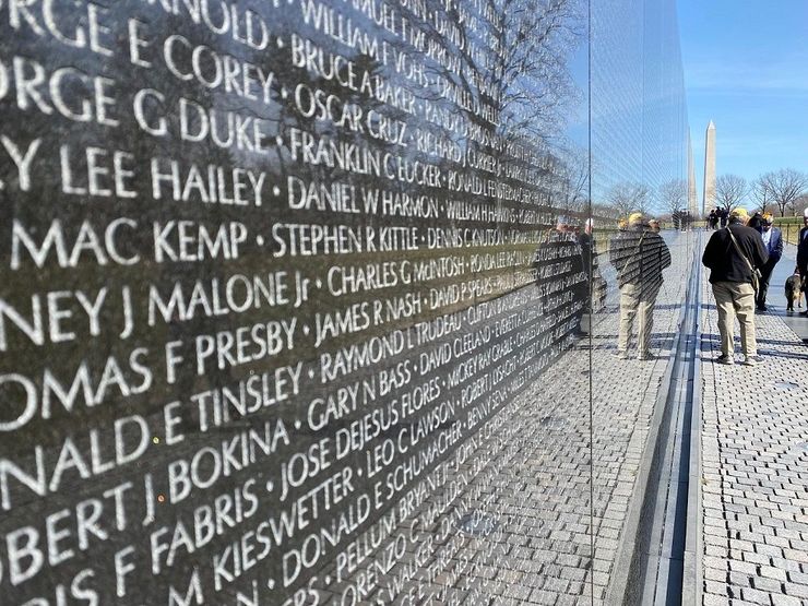 Vietnam Veterans Memorial with engraved names. People are viewing the wall. Washington Monument in background.