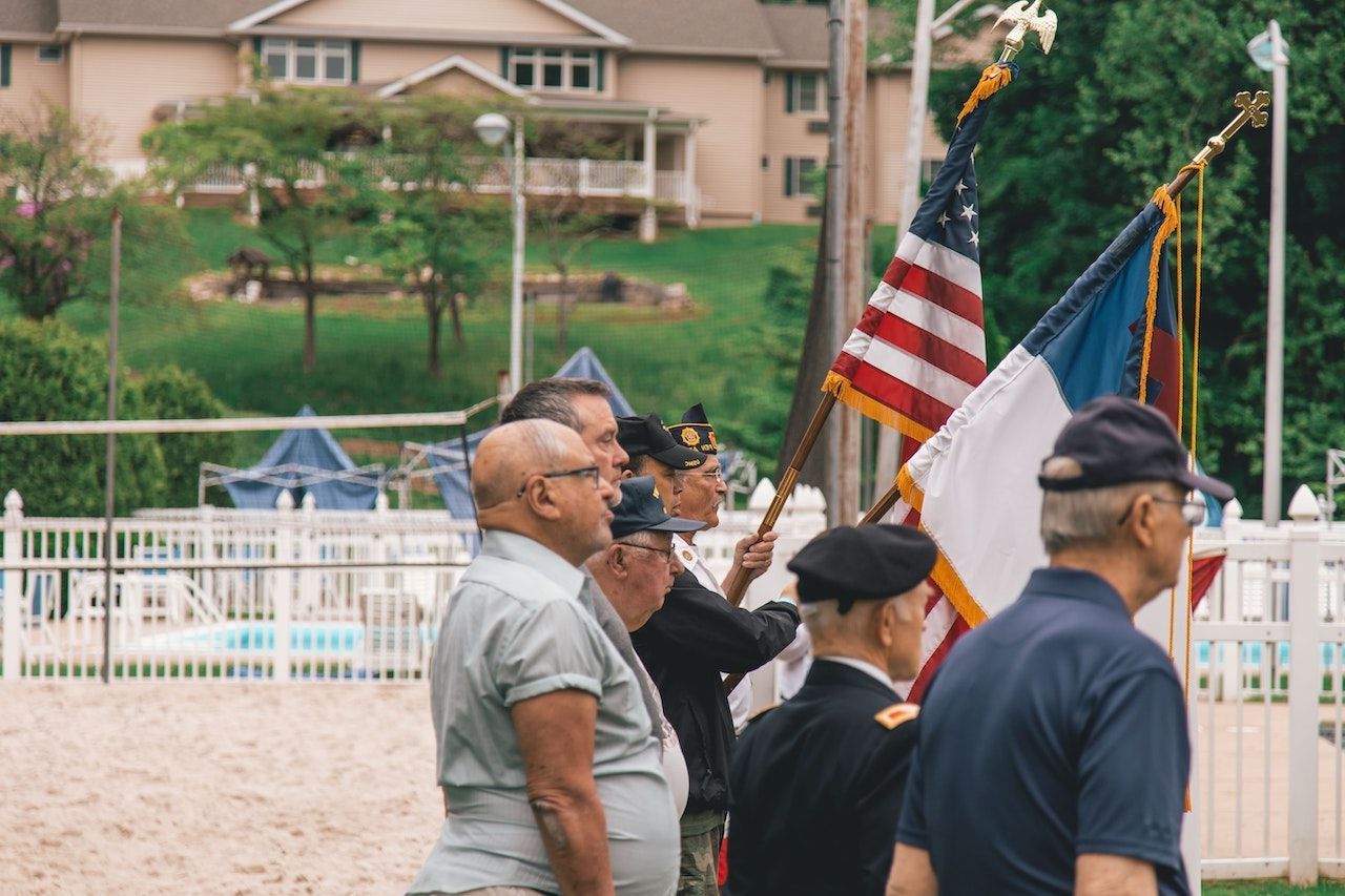 Men holding flags stand outdoors; American, military, and others. A house is visible in the background.