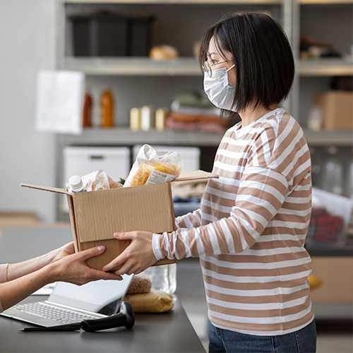 Woman in mask hands a box of food to a person at a counter in a storage room.