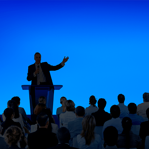 Man speaking at a podium, gesturing to audience in a brightly lit auditorium.