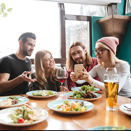 Four people at a table in a restaurant look at a phone. Plates of food and drinks are visible.