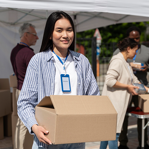 Woman holding cardboard box, smiling, wearing volunteer badge. Others pack items at outdoor event.