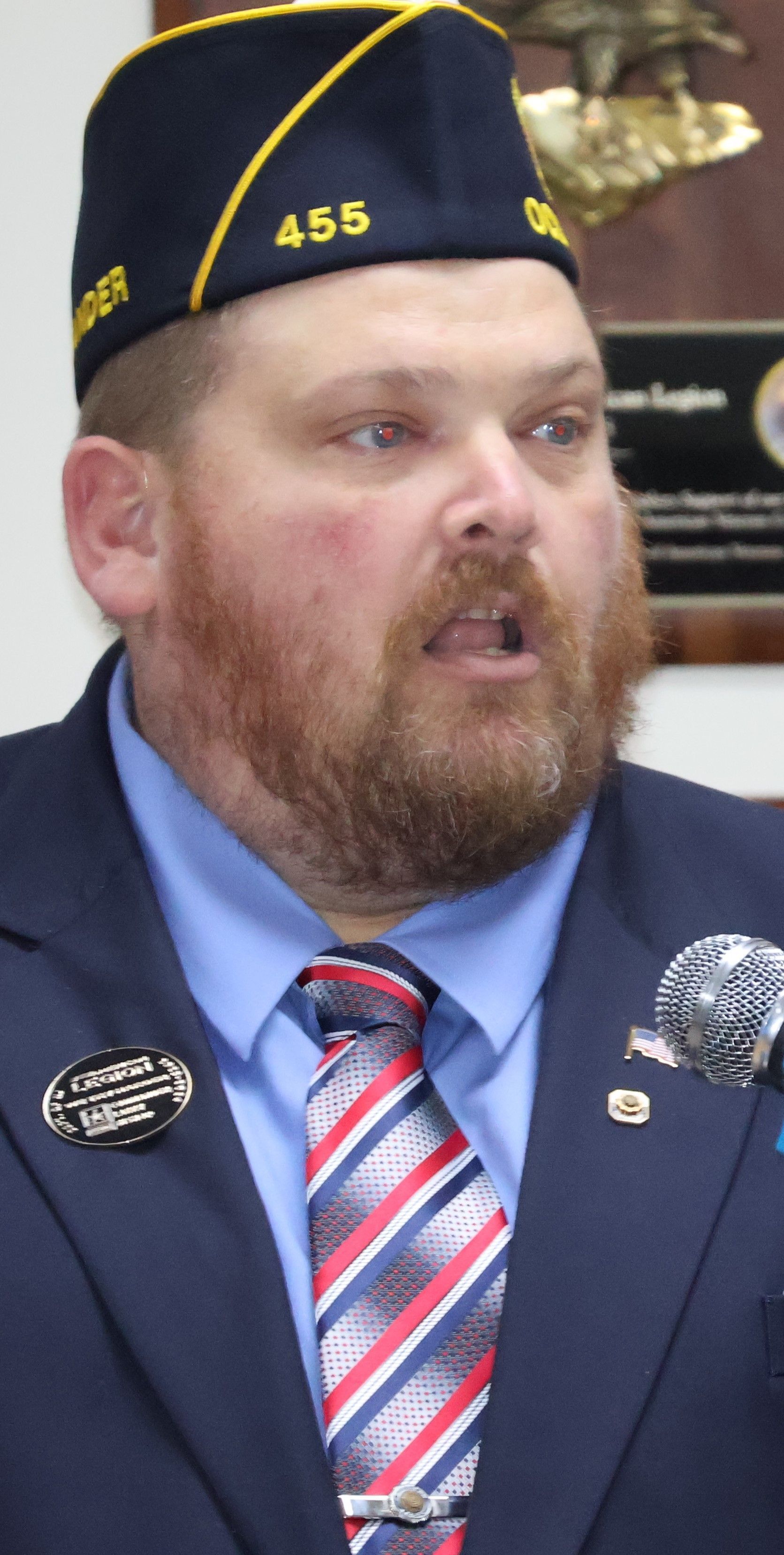 Man wearing a military-style hat and tie speaking, with a microphone and organizational pins visible.