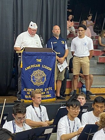 Men at podium with American Legion banner, students in orchestra, spectators in bleachers.