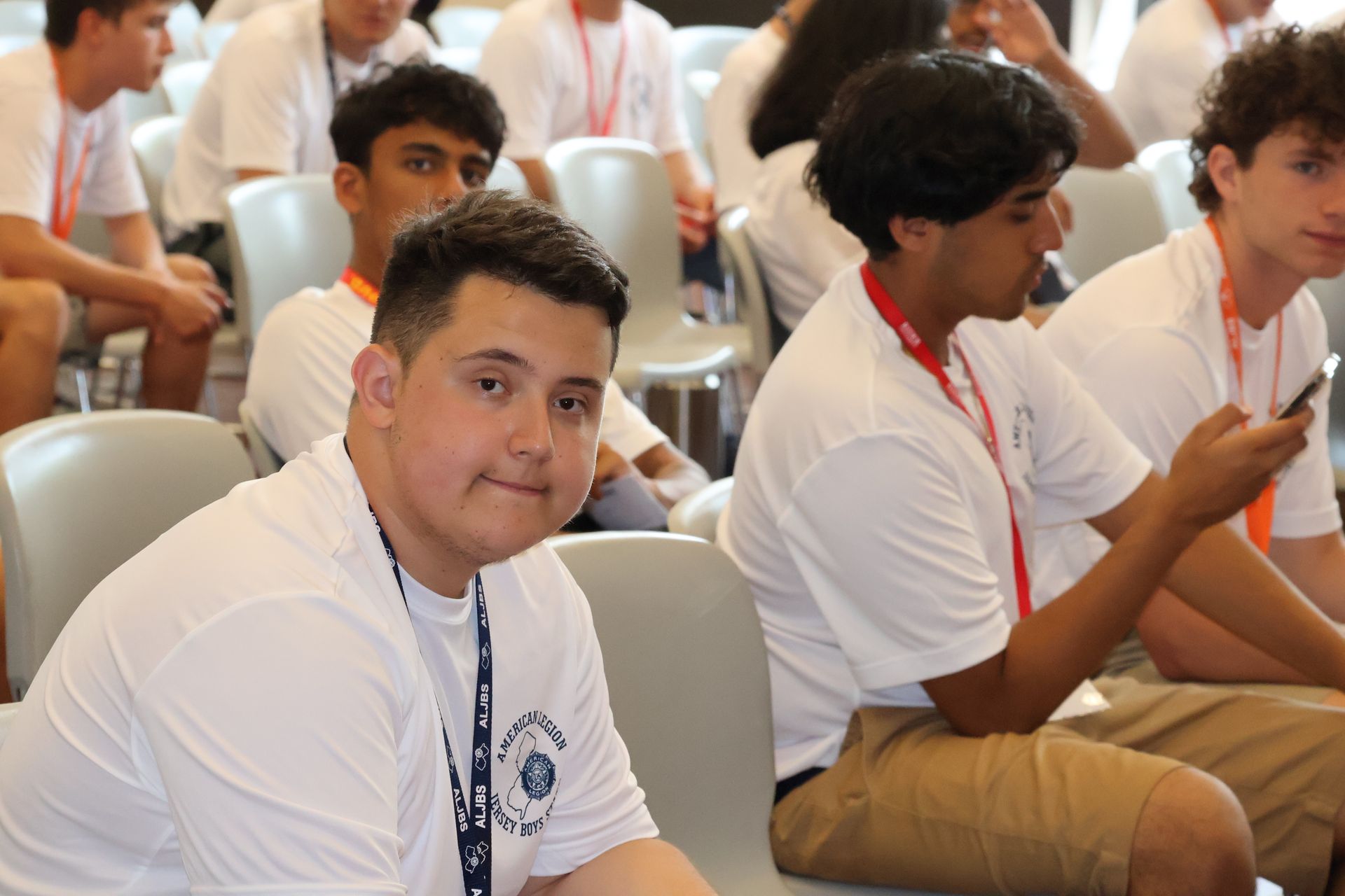 Group of people in white shirts and lanyards sitting in chairs. A person in the foreground smiles at the camera.