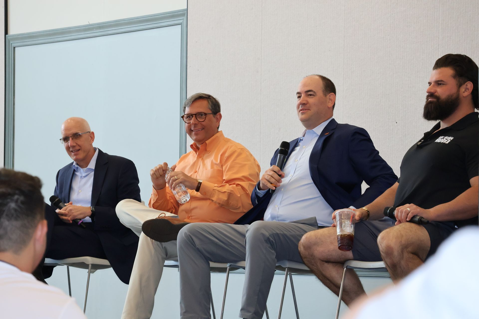 Panel of four men seated, speaking. One holds a microphone, another water bottle. Light-colored wall backdrop.