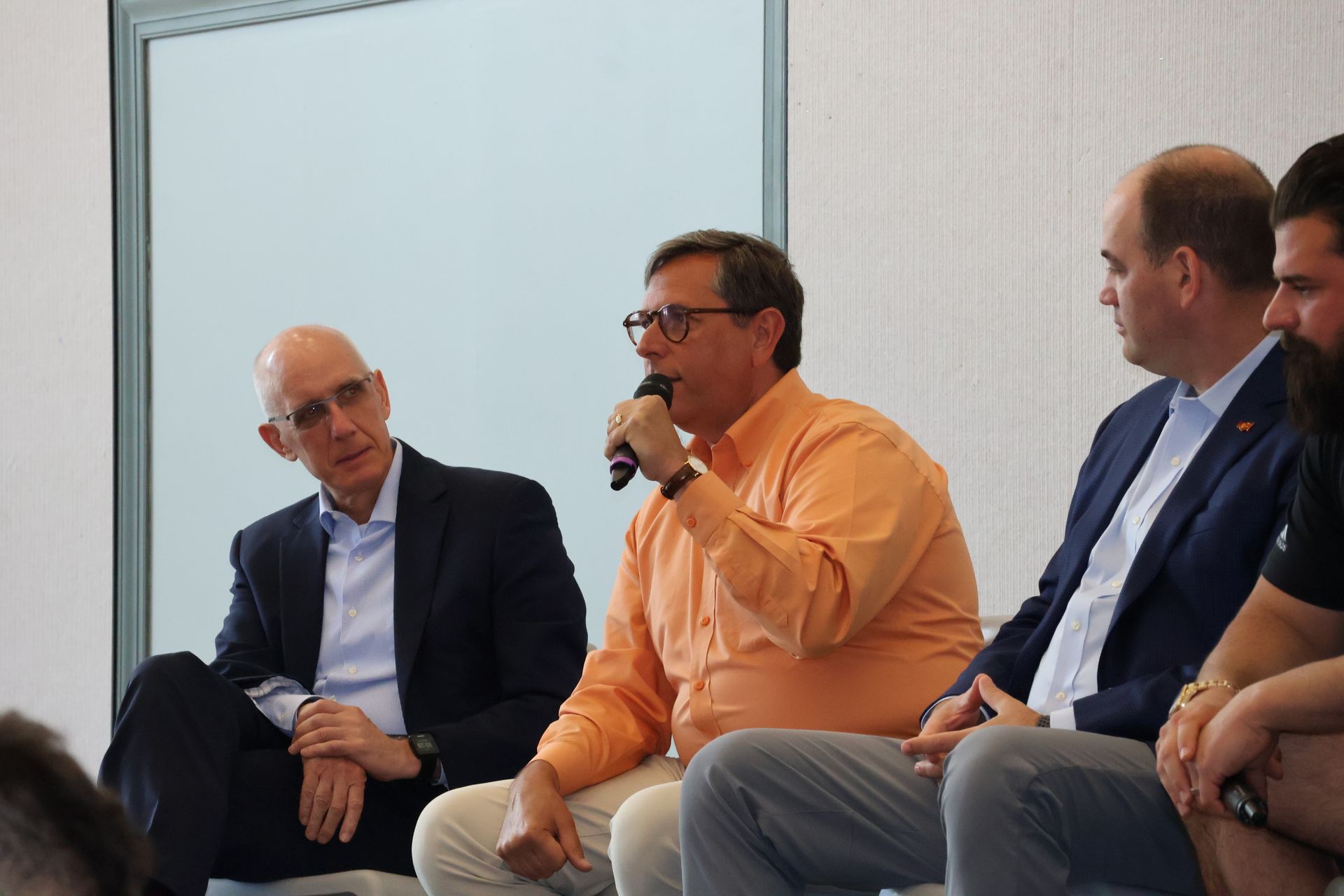 Panel discussion with men. One speaks into a microphone, others listen. Modern setting with white walls.