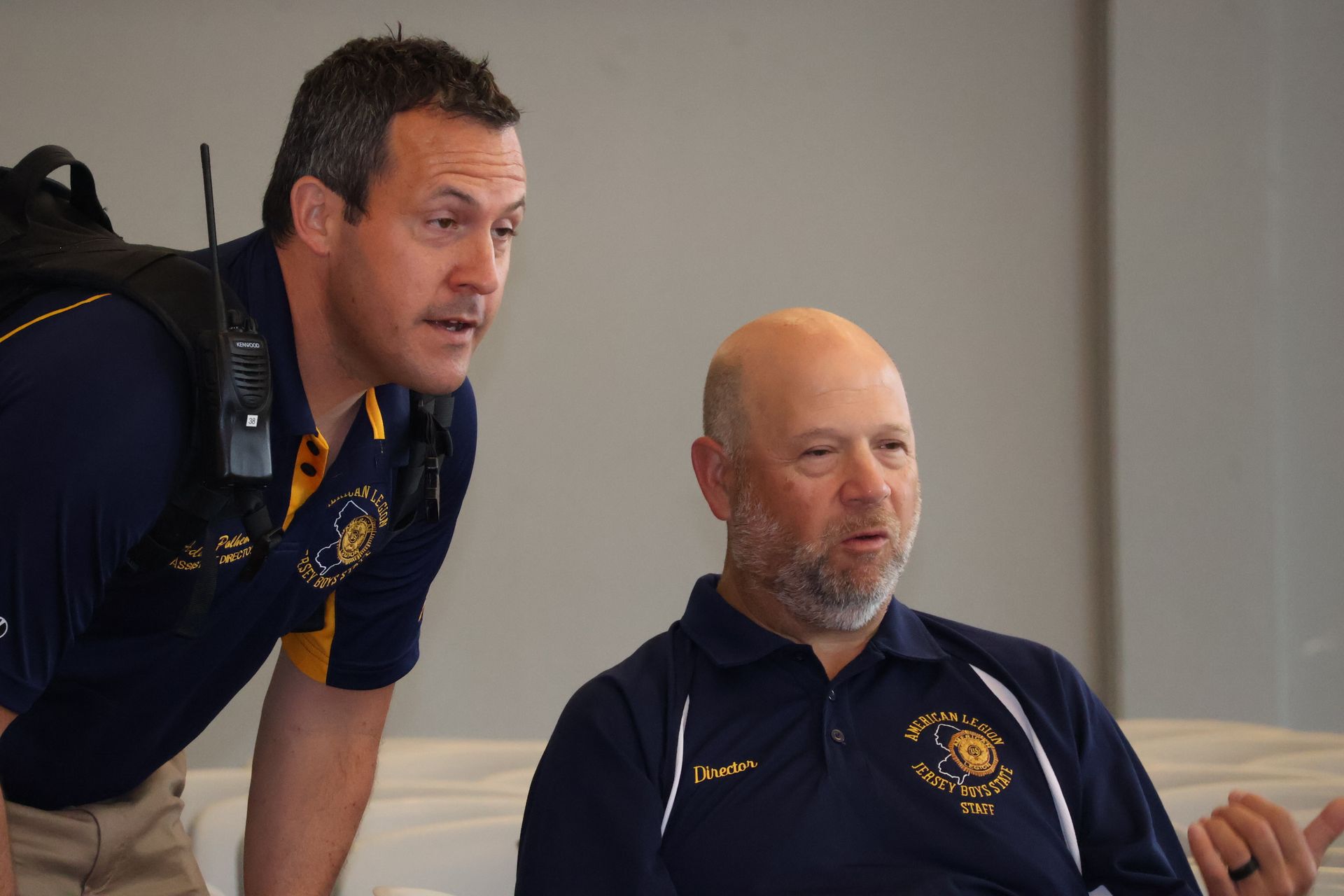 Two men in navy shirts with logos, one leaning in with concern, one gesturing.