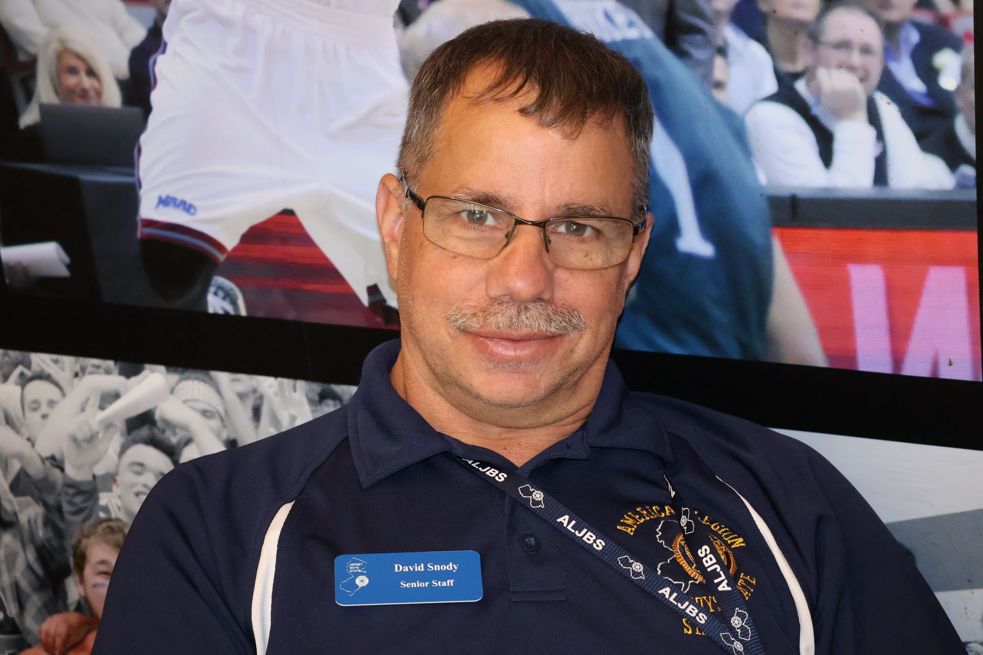 Man wearing glasses and navy polo shirt, smiling, in front of a sports-related background.