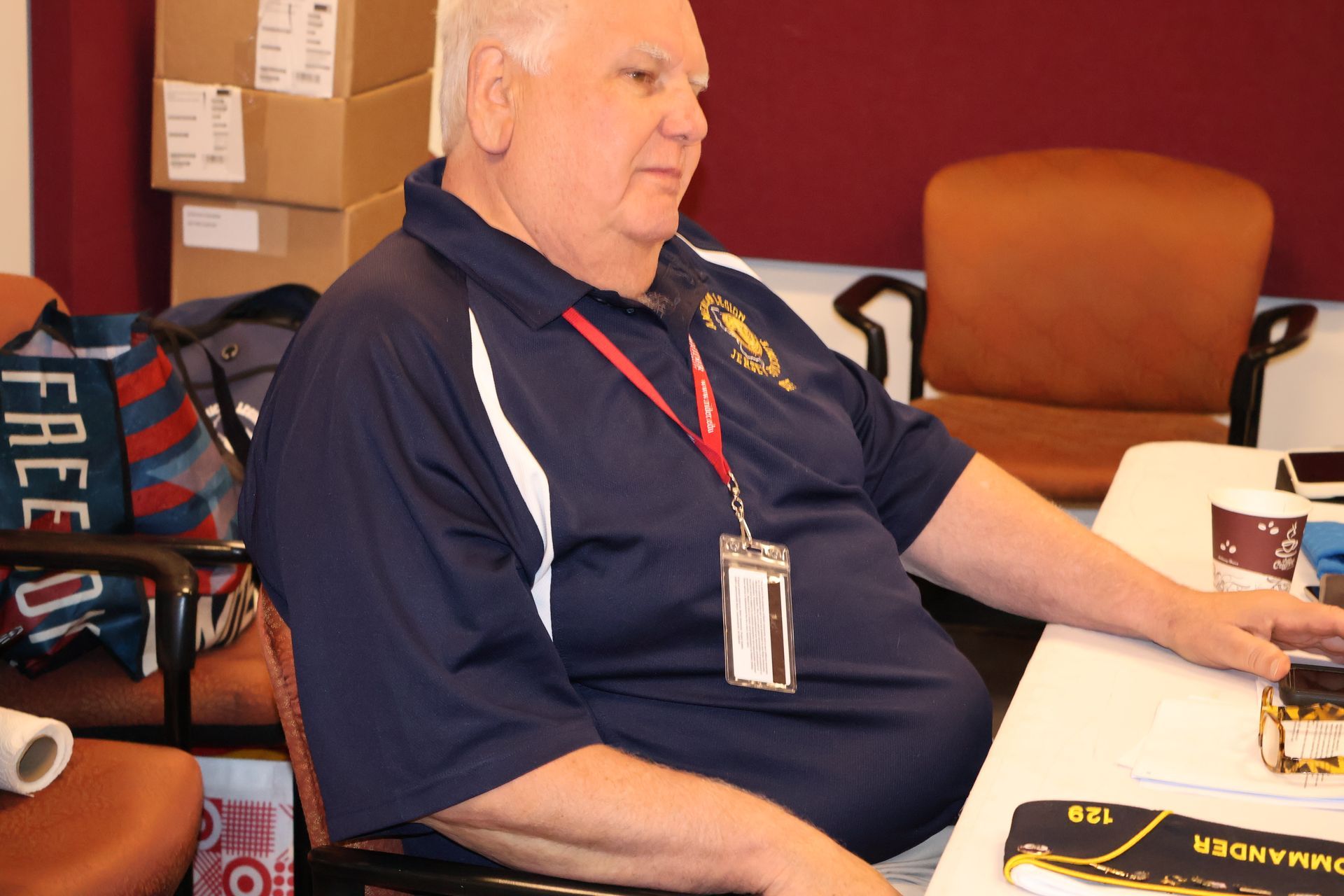 Man in blue polo shirt with lanyard, seated at table, looking to the side.