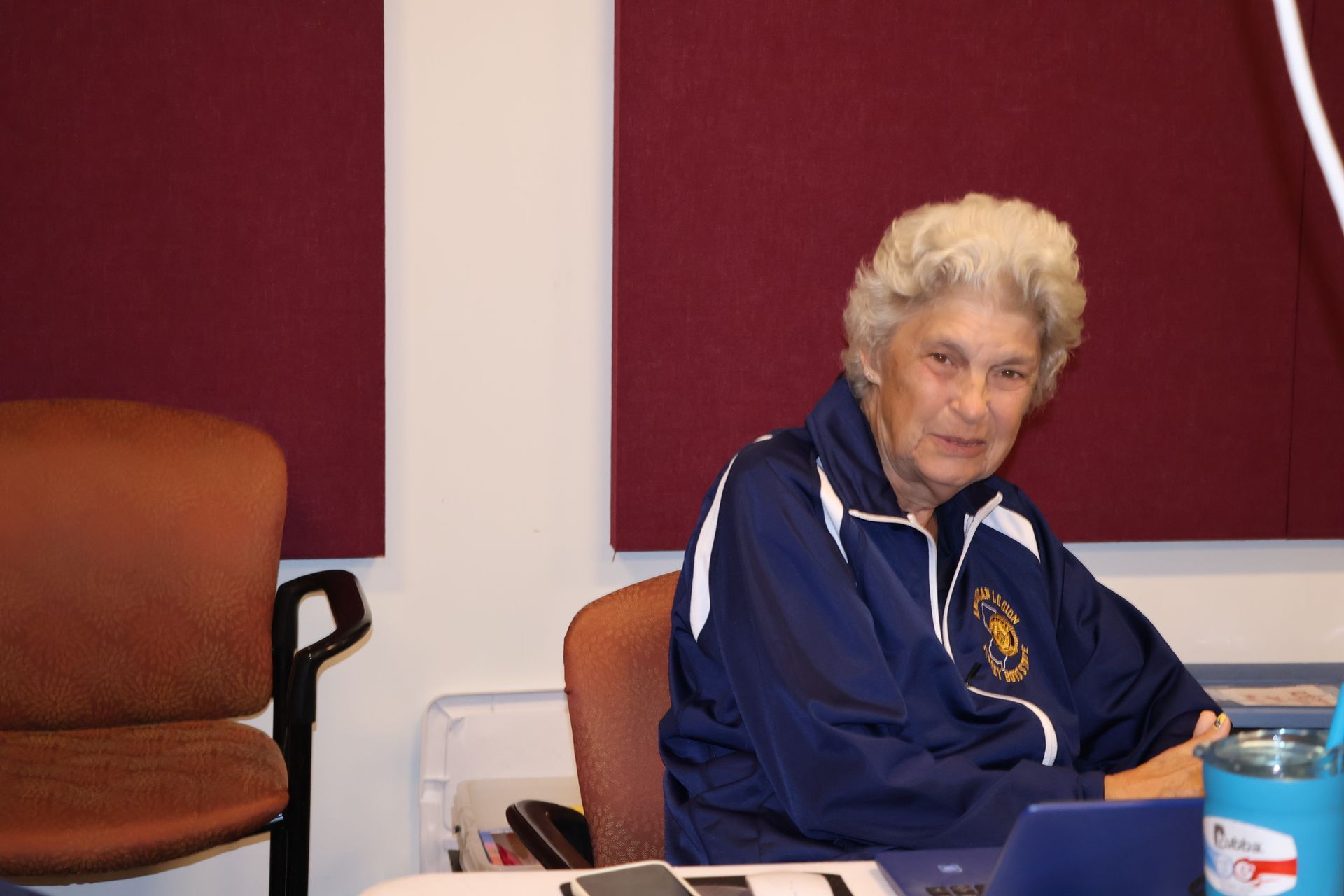 Woman in blue jacket seated at a table, looking at the camera; maroon and white background.