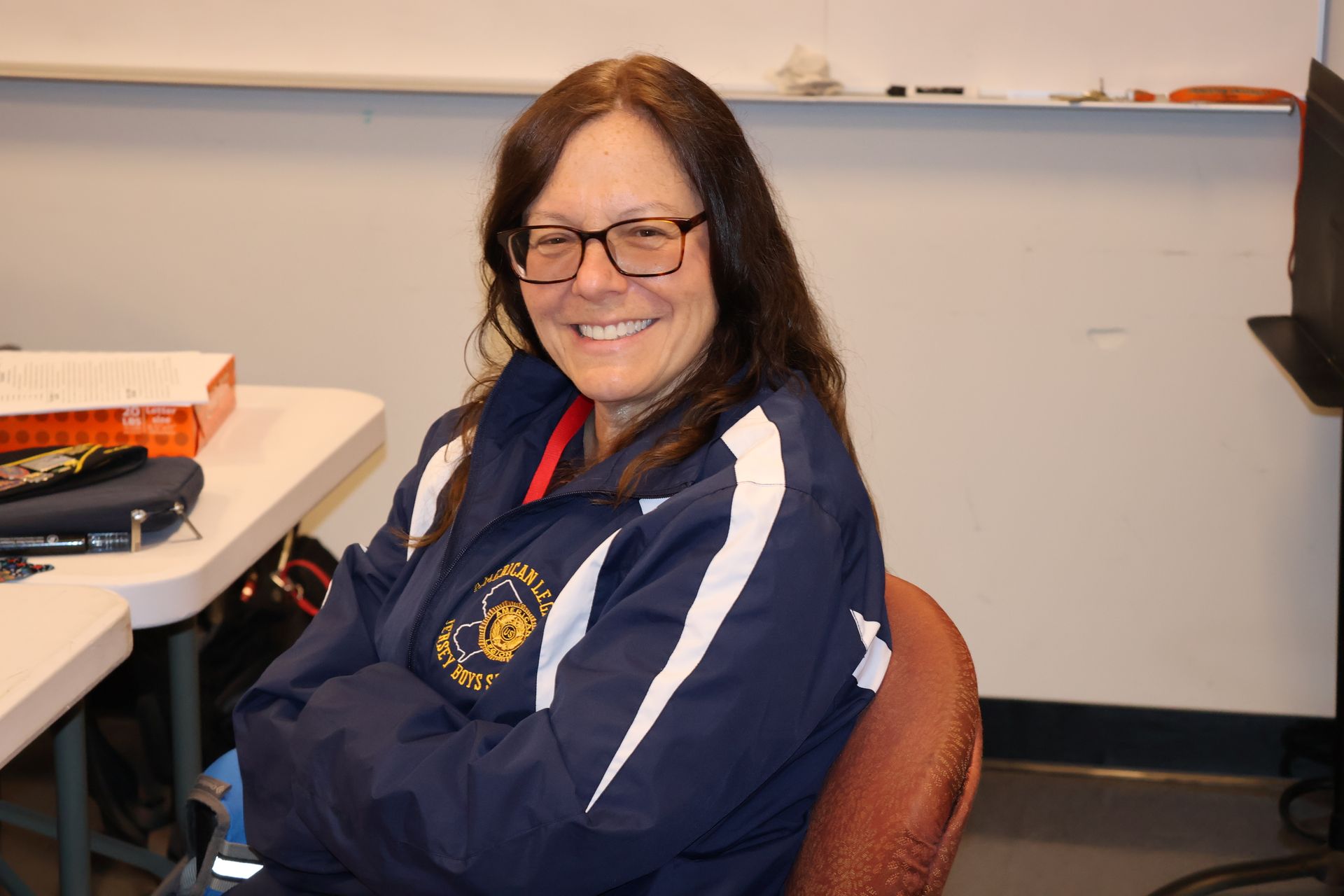 Woman with glasses smiling, arms crossed, wearing a blue jacket, seated at a table.