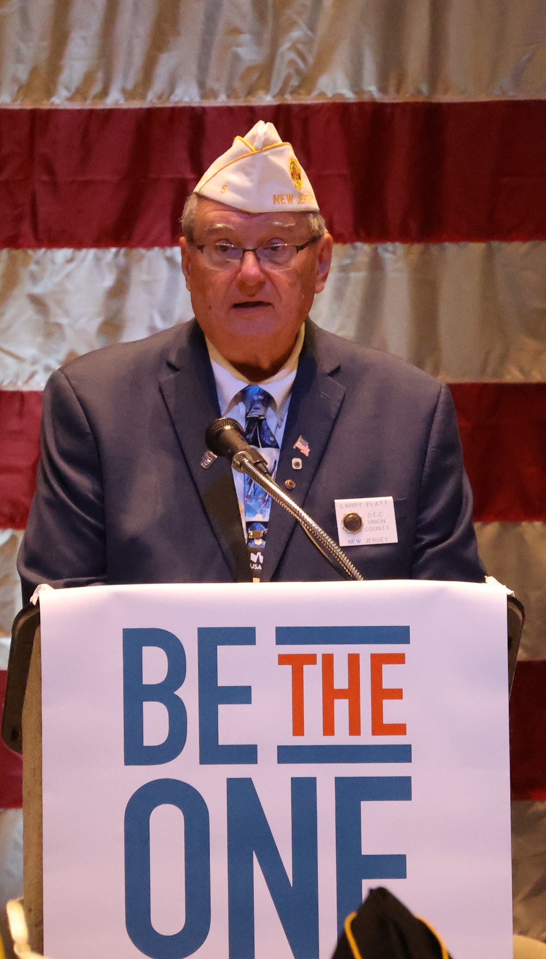 Man speaking at a podium with a “BE THE ONE” sign and an American flag backdrop