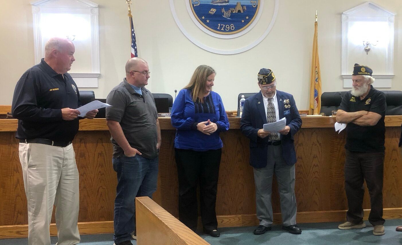 Five people stand behind a wooden barrier, two men hold papers. They appear to be in a government building with flags and a seal.