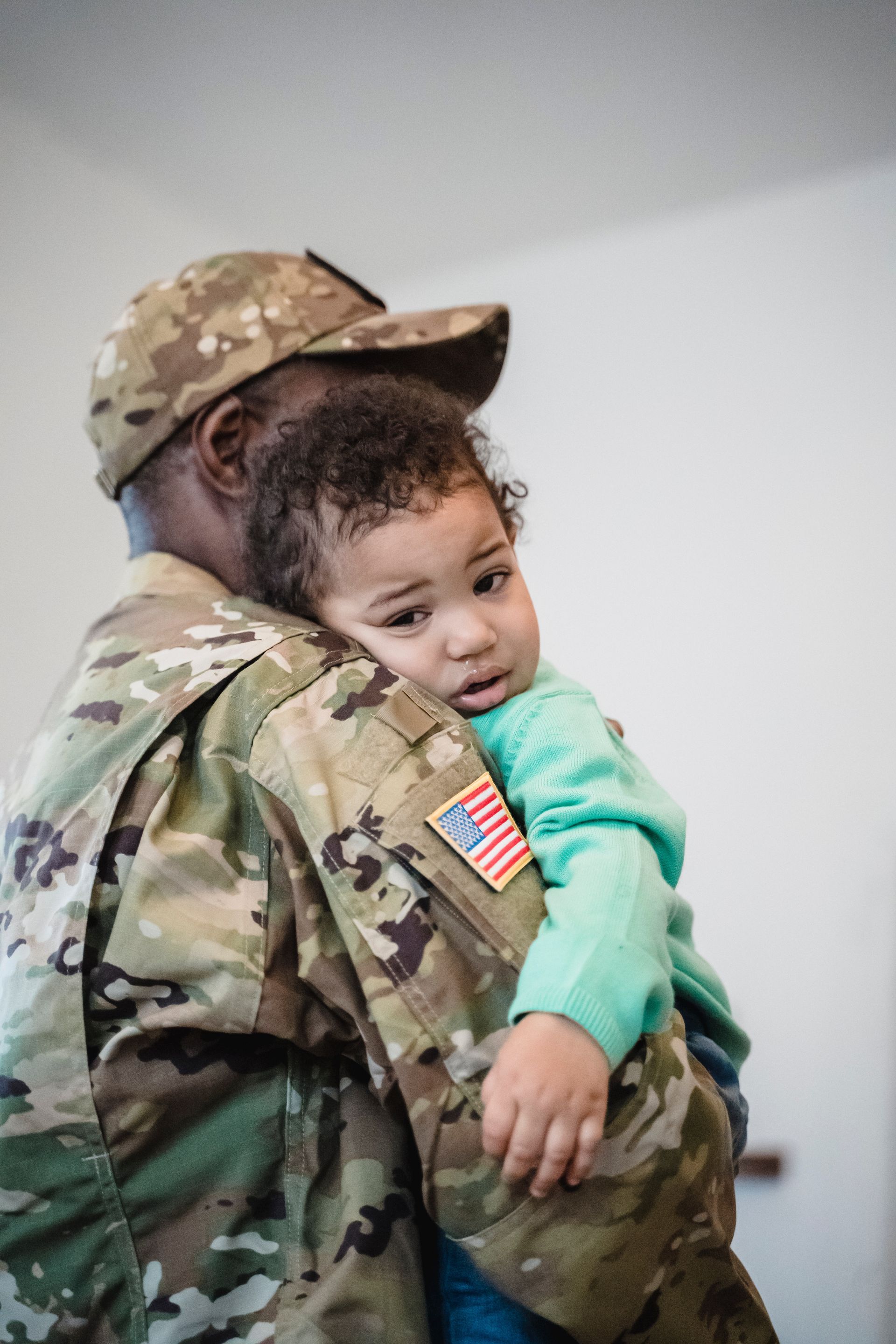 Soldier in camouflage holding a child in a mint green sweater; child looks at the camera.