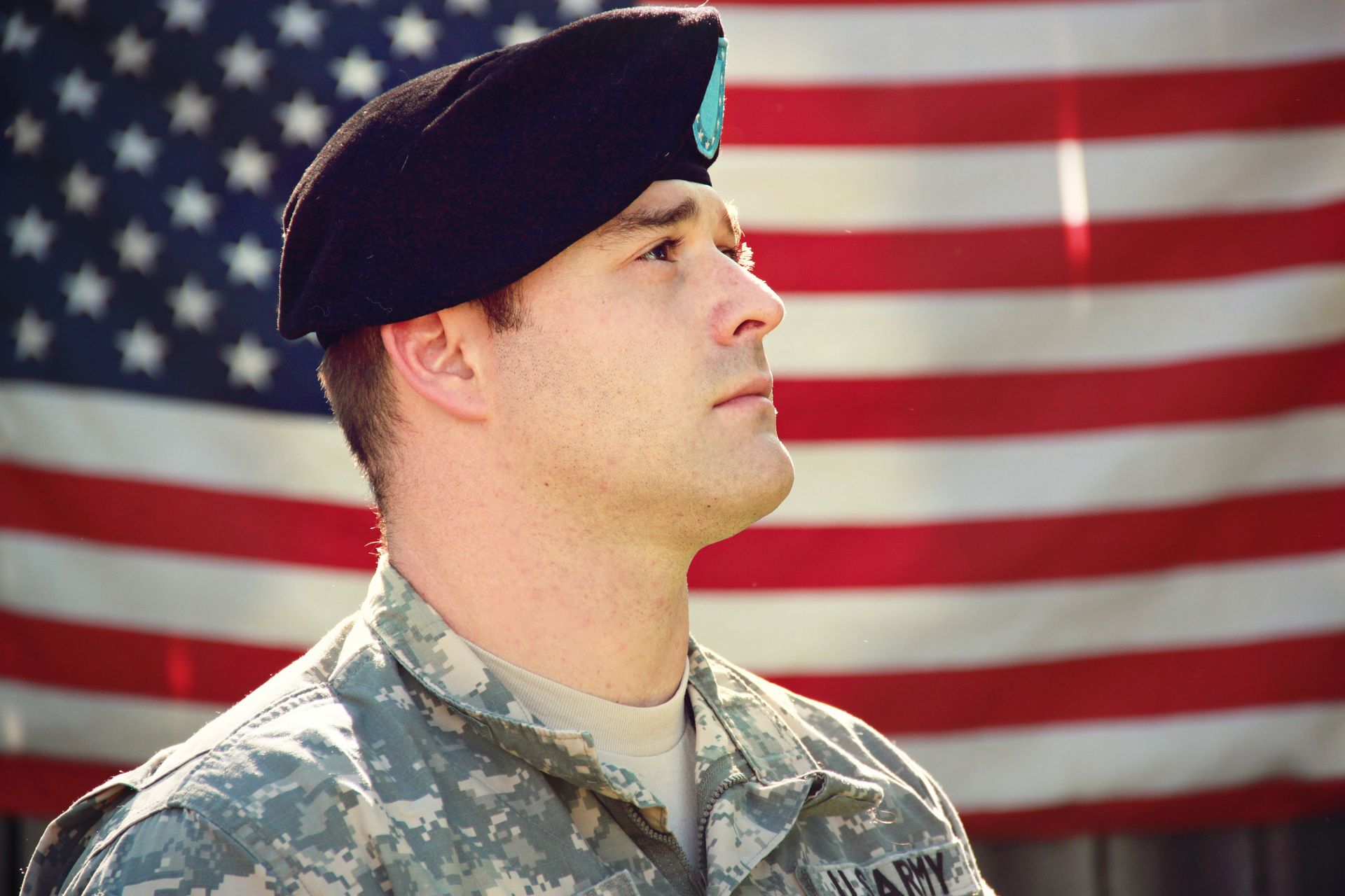 Soldier in uniform and black beret, looks upward, against a blurred American flag.