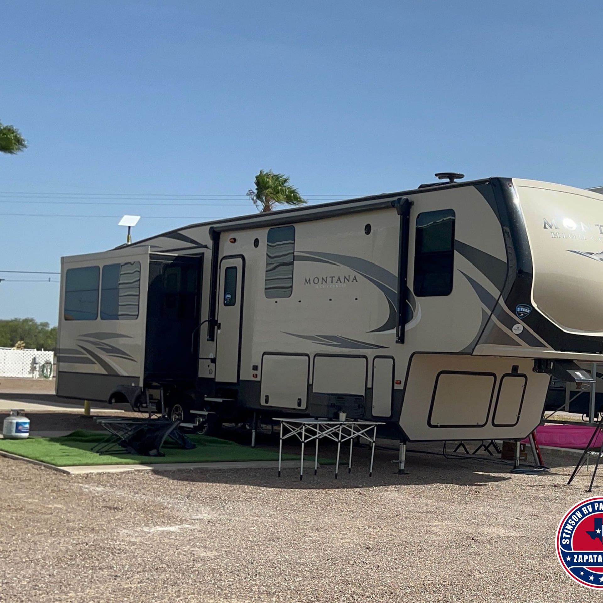 Tan fifth wheel RV parked outdoors on gravel with an open slide-out. Blue sky in background.