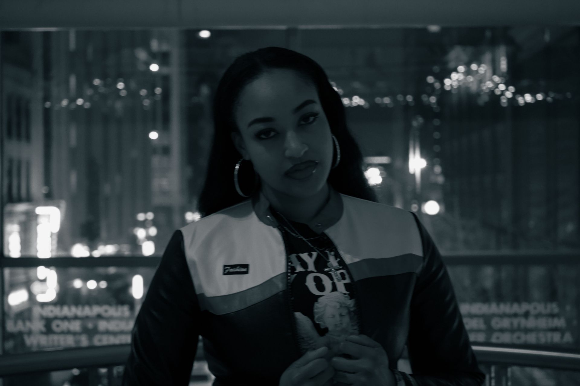 Woman in a jacket holding a teddy bear, standing indoors against a city-light backdrop.
