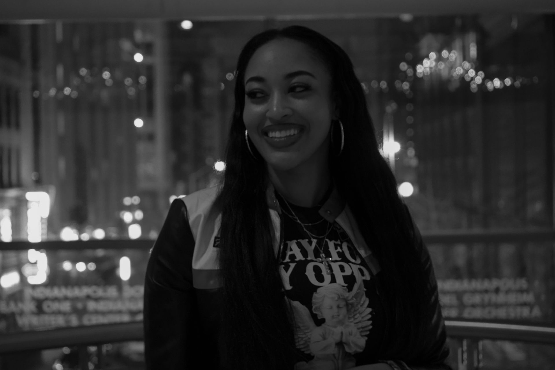 Smiling woman in a dark jacket standing indoors with city lights blurred in the background