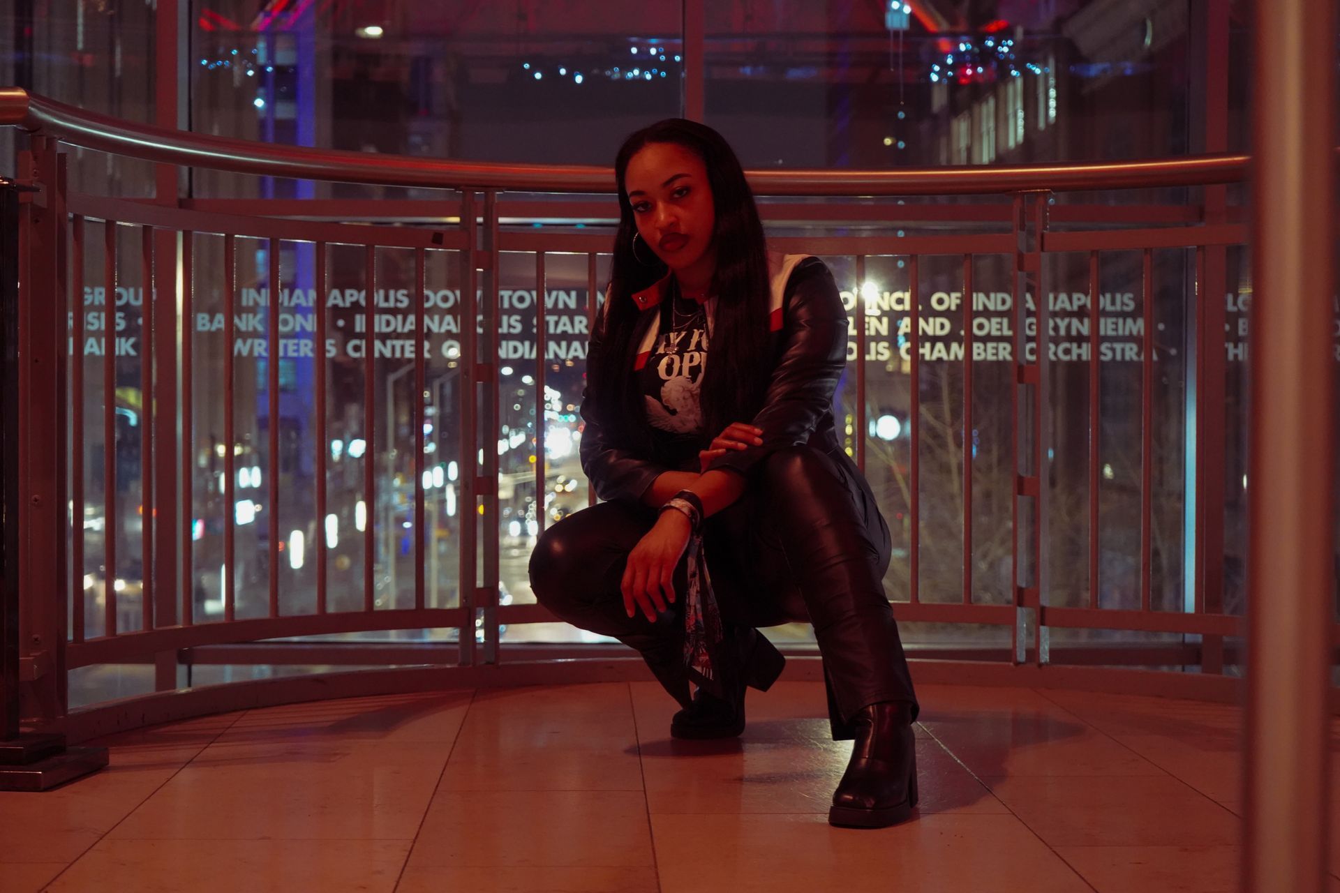 Person crouching on a balcony at night, lit by red neon city lights.