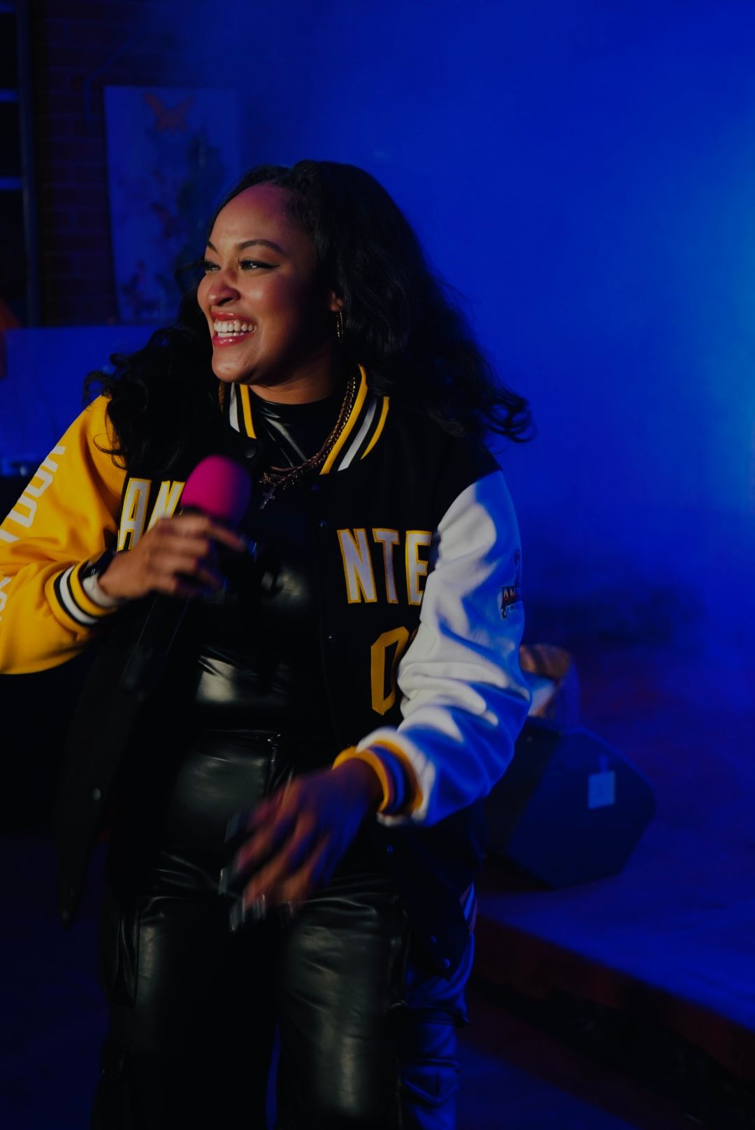 Person singing into a pink microphone on a blue-lit stage, wearing a black and yellow varsity jacket.