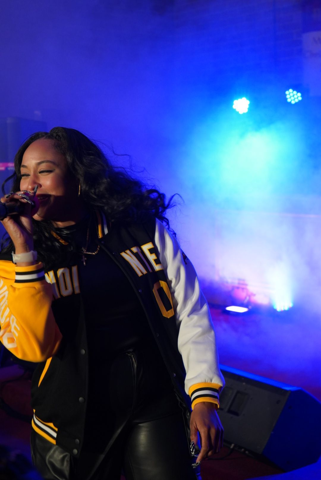 Singer performing onstage with a microphone, blue stage lights and smoke behind her.
