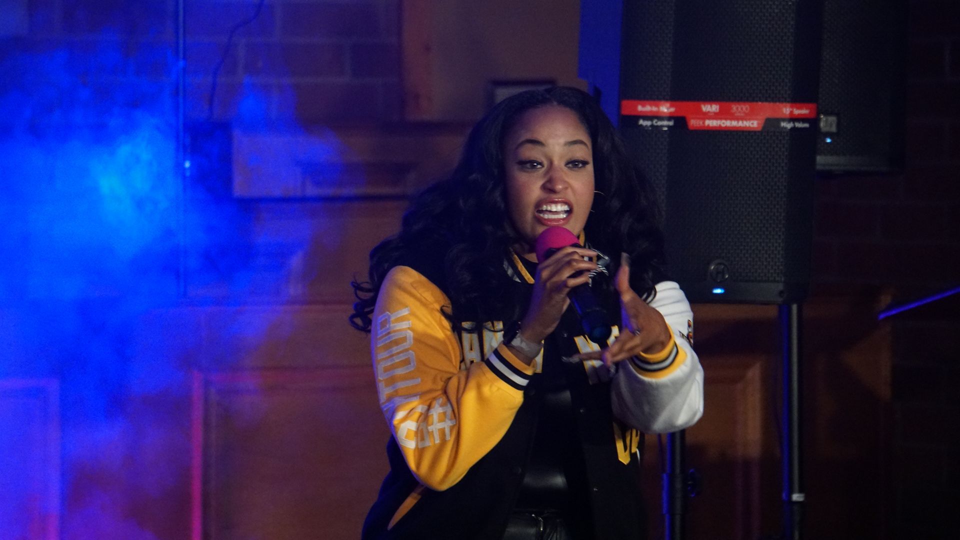 Singer performing onstage with a microphone, wearing a yellow jacket under blue and purple lights