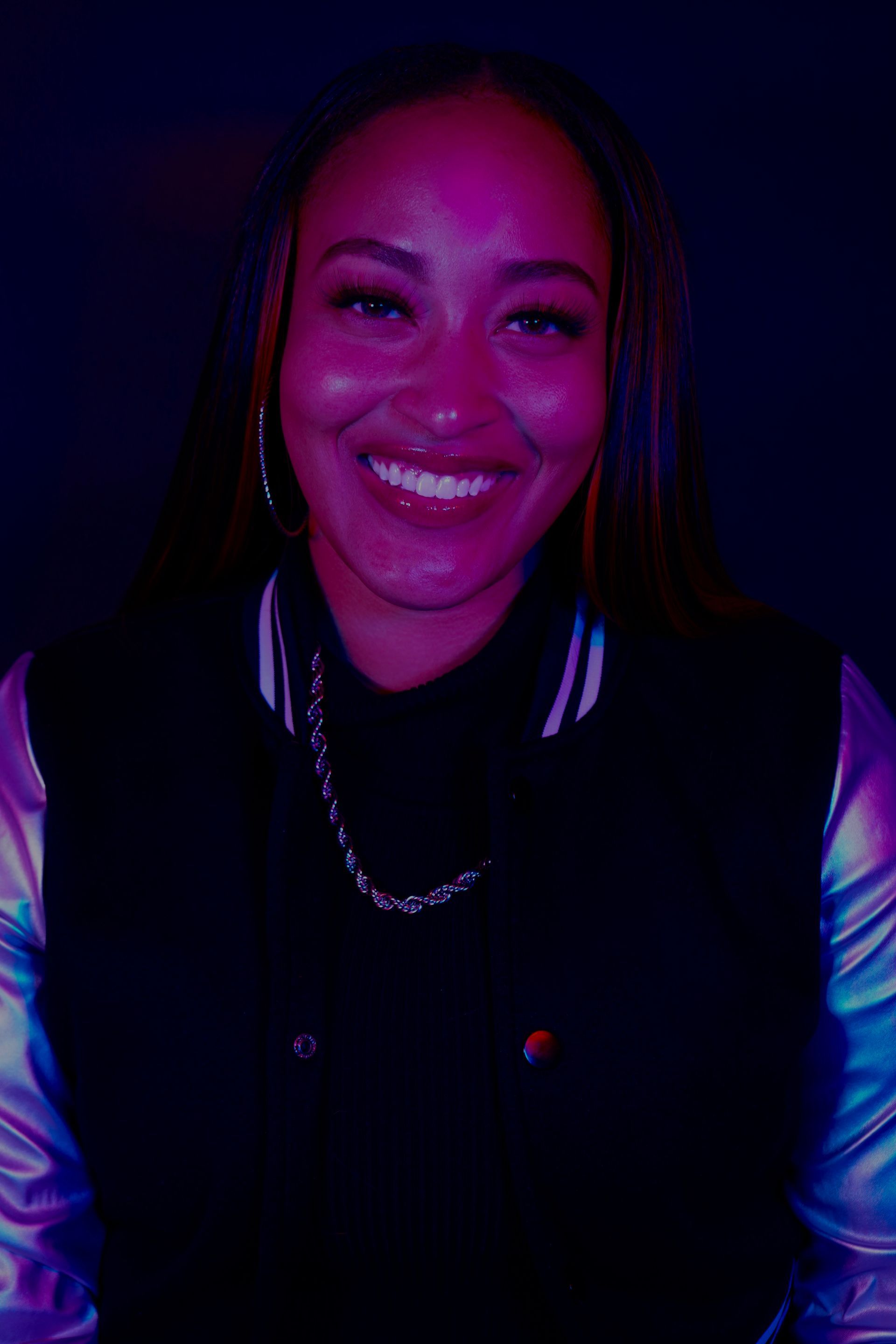 Smiling person in a dark jacket, lit by vivid purple and blue neon light.