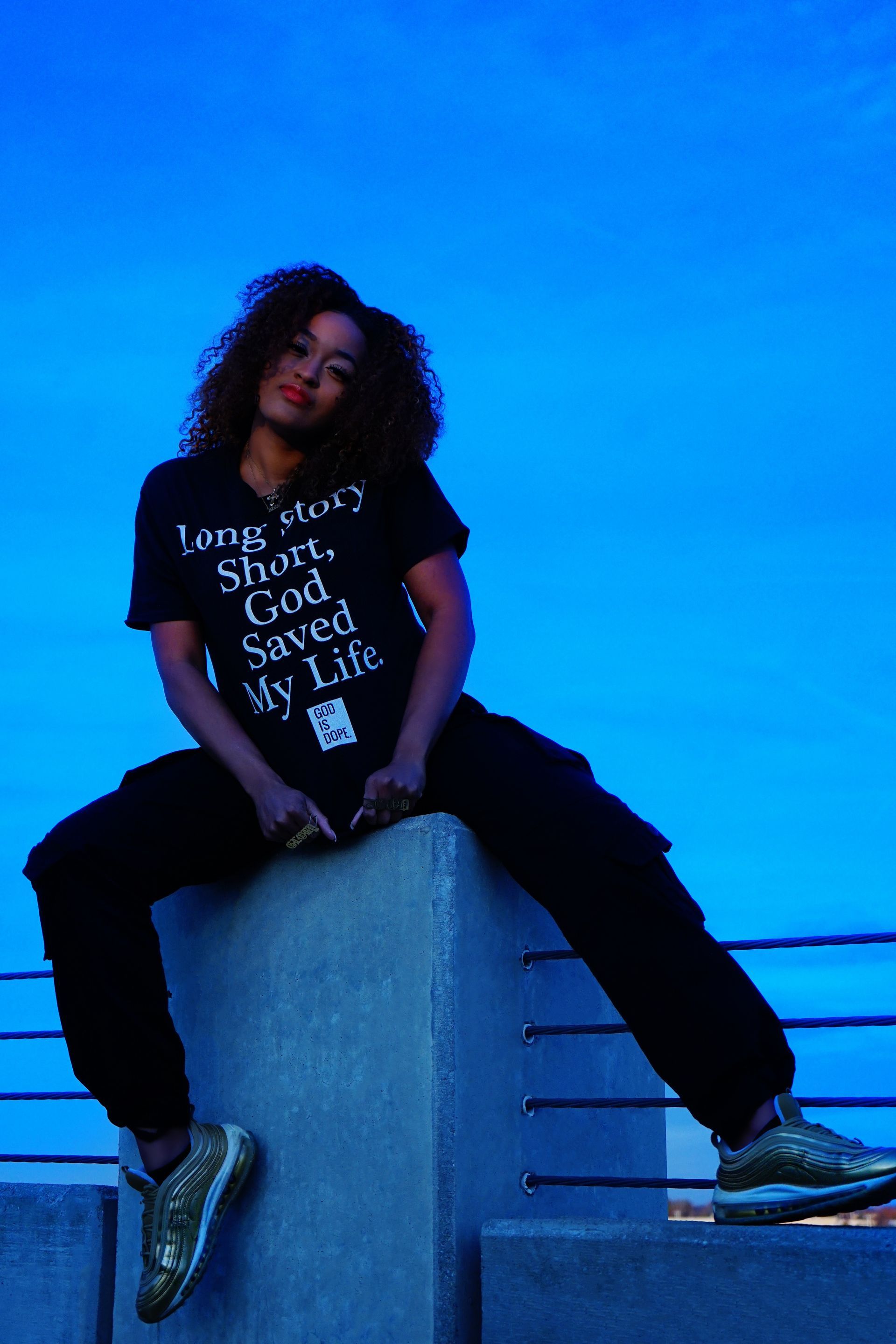 Person sitting on a concrete block against a blue sky, wearing a black shirt and sneakers.