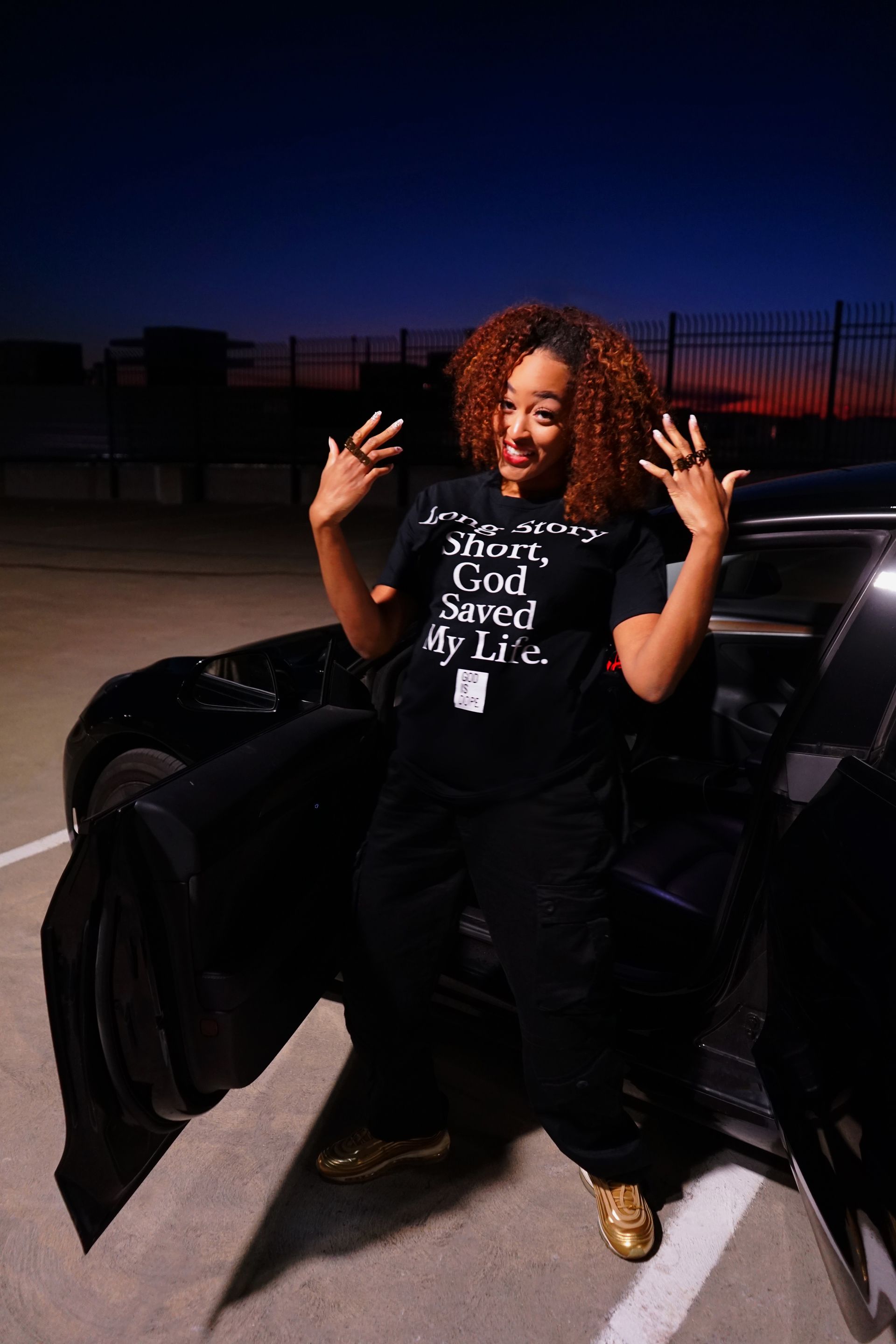 Person posing beside a black car at dusk, wearing a black outfit and gold shoes.
