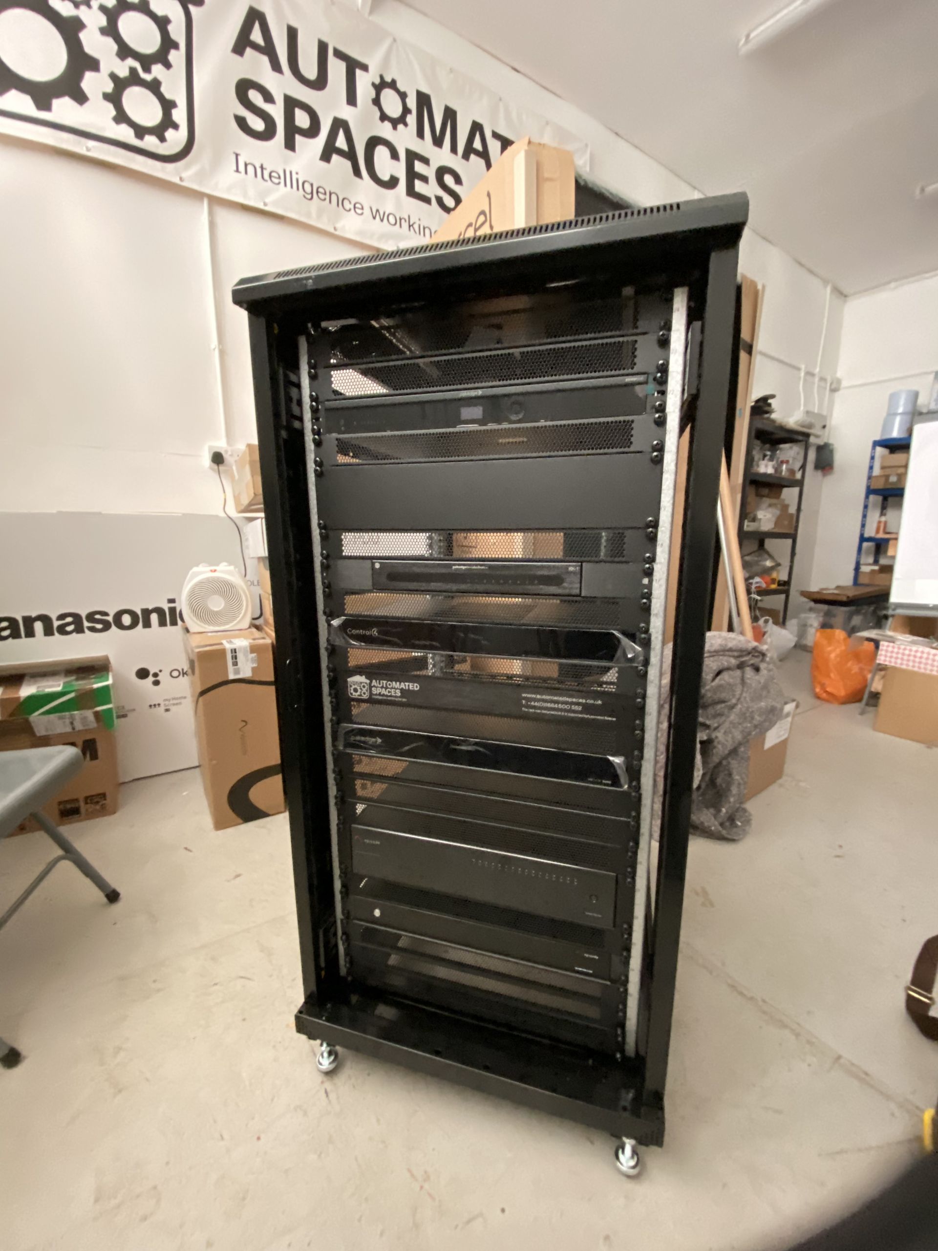 Black server rack on wheels inside a warehouse. Silver rails, shelves visible. Sign for automation spaces.