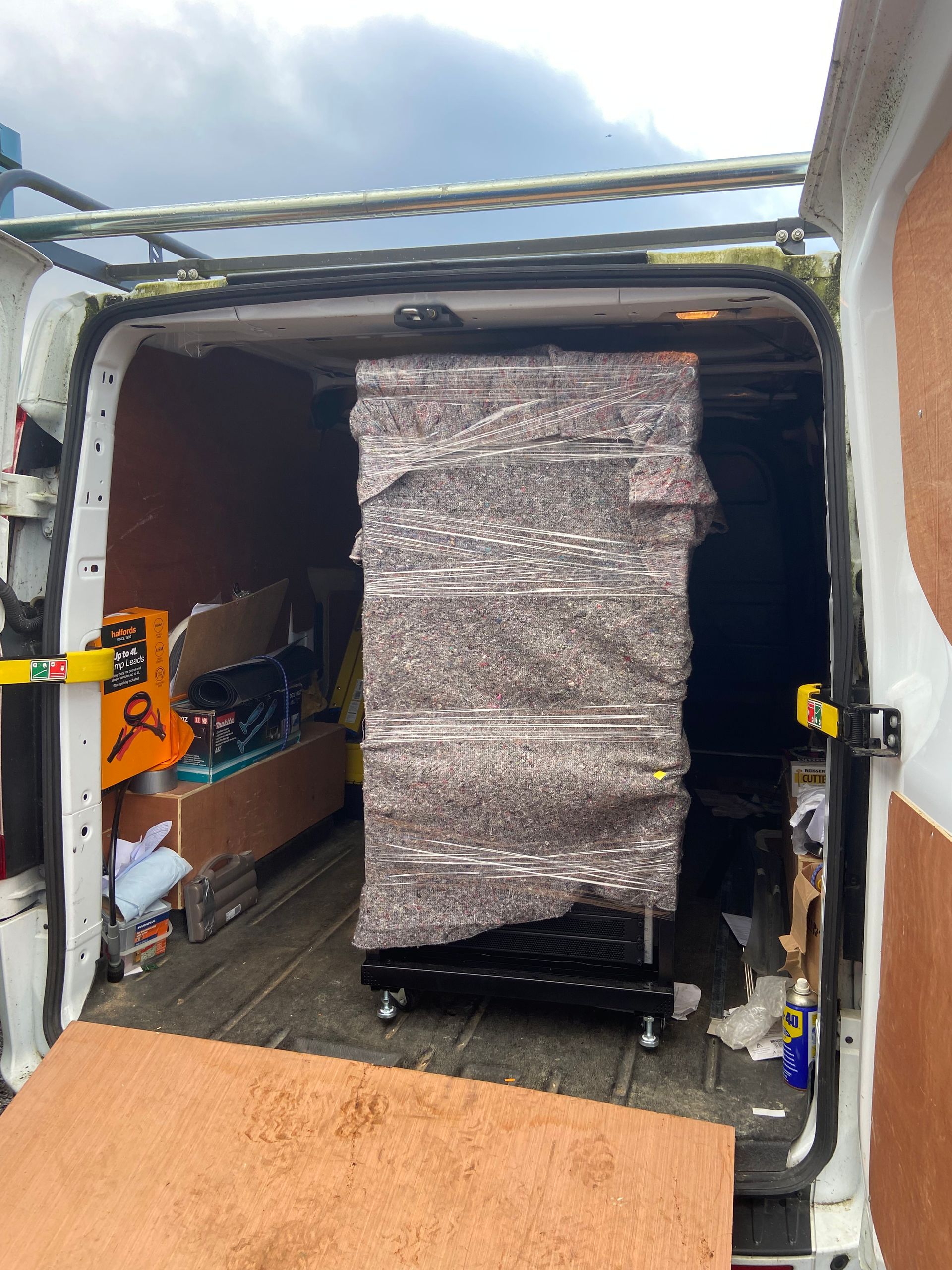 Inside a van: a large, wrapped black object on a wheeled base. The van's doors are open.