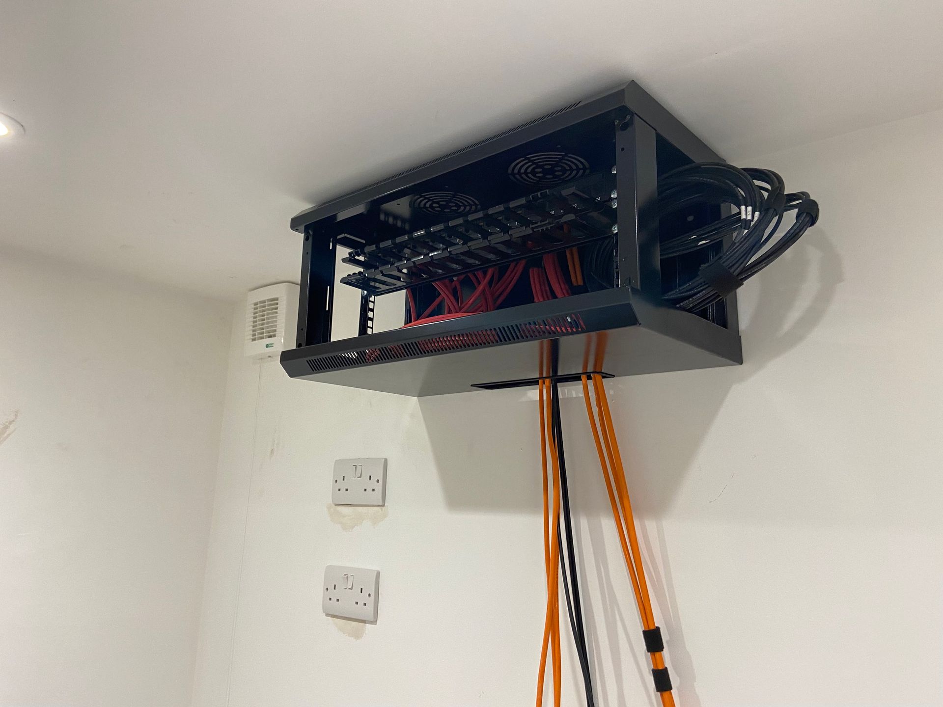 Black patch panel box mounted on a white wall, with orange cables dangling below. Wood flooring.